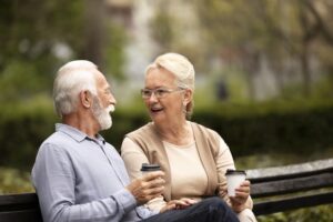 Happy senior couple sitting on a bench in a park, holding coffee cups and talking, symbolizing active and connected living.